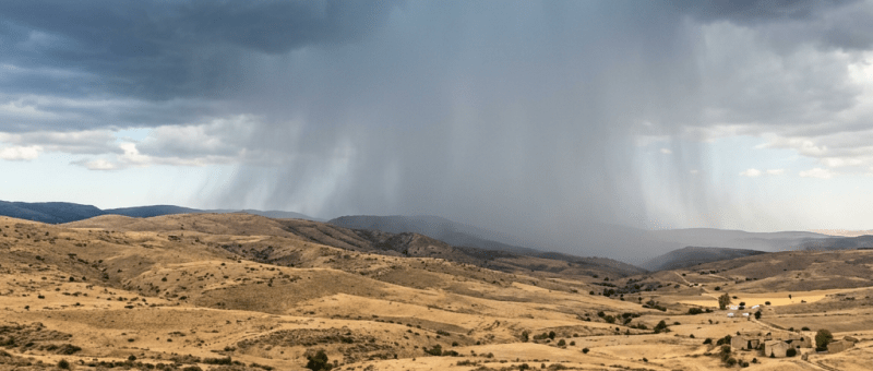 Dark storm clouds releasing rain over dry, rolling hills